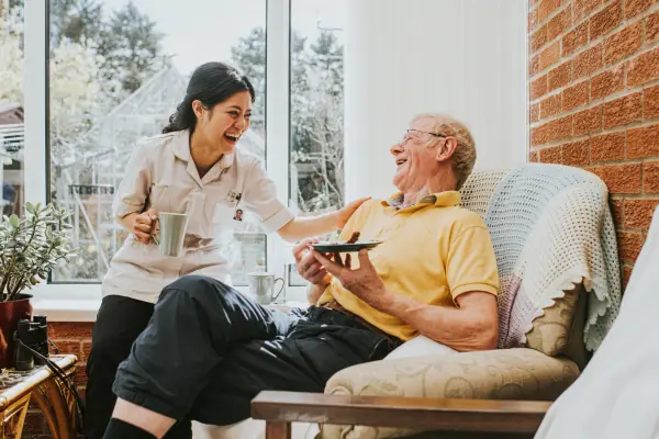 Assisted living caregiver laughing with a senior resident while serving tea in a bright sunroom